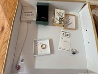 Top down photo showing five pieces of jewelry arranged in an open box, includes necklaces in plastic bags and rings in small boxes or loose.