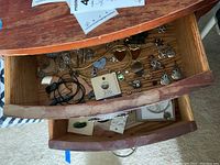 View into a wooden drawer showing assorted jewelry pieces like earrings, lockets, pins, and necklaces scattered inside along with a round jewelry box and a heart-shaped box.