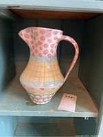 Front view of the 10 inch ceramic pitcher with pink polka dots and yellow grid pattern on a blue shelf.