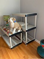 Front angled view of the plastic shelving unit assembled in two pieces with five shelves total, placed against a white wall on wood floor. Includes objects not part of sale.