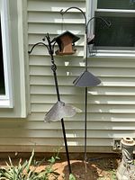 Two shepherd's hooks side by side, each with bird feeders and metal baffles, set against beige vinyl siding with some garden plants at base.