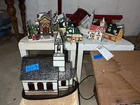 Wide photo showing one large stained glass church lamp with a black roof, white wall panels, brown window panes, placed near eight smaller colorful holiday houses on a white table and the floor in a basement setting.