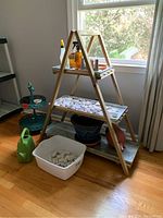Photo showing wooden three tiered plant stand with metal trays, garden stones filling trays and plastic tub, green plastic watering can, green two tiered metal stand and plant pots nearby
