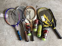 Five tennis racquets lying on concrete floor, each with different colored handles, showing wear. Four cans of Wilson tennis balls placed in front. Two racquet covers are beside them.