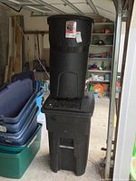 Stacked black plastic trash cans with lids on wheels, Rubbermaid Roughneck 45 gal. on top, Toter 64 gal. below. The garage setting with plastic bins visible in background.