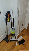 Photo of two cordless vacuums and a white steam mop with accessories, standing against a wall with hardwood flooring visible.
