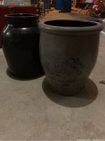 Two vintage crocks, one dark brown and one gray with blue writing, placed side by side on a basement floor.
