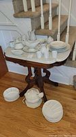 Photo of vintage marble top table with multiple white ceramic dinnerware items arranged on and around it, showing table and contents from front and side angles.