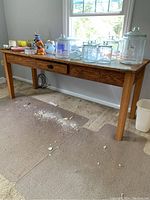 Full view of the vintage solid wood work table against a wall underneath a window, showing the single drawer, various glass jars and containers placed on top, and the clear plastic chair mats underneath the table.