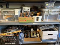 Multiple plastic bins and cardboard trays on metal shelving containing battery packs and loose cells