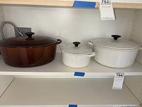 Front view showing all three Dutch ovens on shelf: brown oval, white small round, white large round