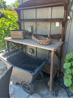 Wooden potting bench with attached top shelf, two wicker baskets on bench, black plastic wheelbarrow underneath