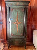 Front view of the media armoire showing the painted green door with central compass star inlay, decorative animal and floral borders, and bottom drawer with knobs.