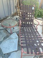 Outdoor heavy metal lounge chair with reddish-brown woven metal straps and curved armrests next to round glass-top side table on stone and gravel ground.