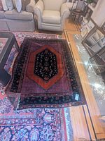Oblique view of rug laid over another larger rug, shows overall pattern, red field, central medallion, floral border and surrounding furniture