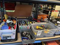 Three clear plastic bins and one gray basket on a shelf filled with various hand tools and hardware