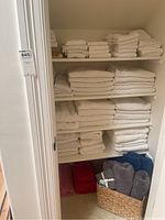 Image of three shelves filled with white towels of various sizes, along with a basket on the floor containing gray and dark blue towels, and folded red towels underneath the basket