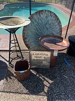 Overview of metal wall art, birdbath on stand, square pottery planter, two ceramic pots, and a garden sign grouped near pool