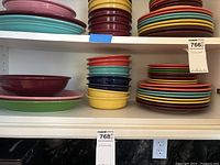 Shelf with stacks of colorful Fiestaware dinner plates, salad plates, and bowls including pastel and bright shades like red, pink, yellow, turquoise, navy blue, green, and orange.