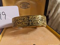 Close-up of bracelet showing ornate niello design and gold-filled metal finish with jeweler's box logo visible inside.