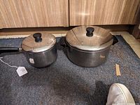 Both Revere Ware stainless clad cooking pans with their glass lids and black handles shown together on a carpet.