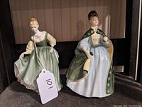 Photo showing two Royal Doulton porcelain figurines on display, one in a green dress and the other in white dress with green cloak