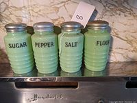 Front view of four pale green Jadeite glass shakers labeled SUGAR, PEPPER, SALT, FLOUR.