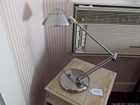 Desk lamp positioned on a wooden side table next to a window air-conditioning unit, showing the arm directly forward and the shade angled downwards.