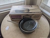 View of the Calphalon 5 quart nonstick saute pan and its lid placed on a table beside the original box.