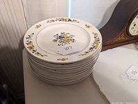 Stack of 12 white ceramic dinner plates with yellow floral pattern around edges and center, showing mostly very good condition with some small chips on a few plates.