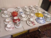 Table display of multiple porcelain teacup sets, saucers and plates in various floral patterns plus a red and gold teapot