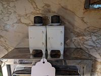 Two white vintage salt and pepper shakers in the shape of monitor top refrigerators with metal screw tops, placed on a reflective kitchen scale.
