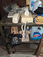 View of multiple old telephones including rotary dial and push-button models on an industrial shelving unit, some with coiled cords and wired handsets.