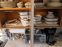 Wide view of kitchen cabinet showing various sizes of CorningWare casserole dishes stacked and coffee pots placed.
