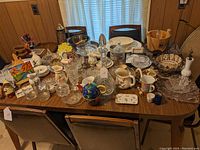 Full view of the large mixed lot showing the range of glassware, ceramics, metalwork, and decorative items arranged on a wooden table with chairs.
