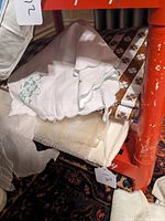 Photo showing white embroidered napkins with scalloped edges, cream textured cloths, and brown and white damask patterned textile pieces stacked beneath a red shelf.