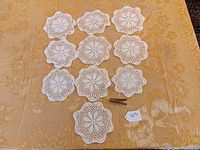 Top view of 10 white crochet doilies/coasters arranged on a gold textured tablecloth. Each piece has a consistent floral pattern and scalloped edges.