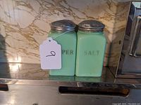 Two jadeite glass shakers with metal lids, stamped 'PEPPER' and 'SALT' in muted letters, placed side by side on a reflective surface with marble background.