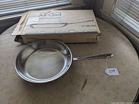 Photo shows the 12-inch stainless steel fry pan with long handle on table next to its original cardboard box and lot tag 229.