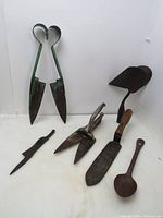 Front view of six vintage garden tools including shears, dirt scraper, pronged tool, garden scissor shears, metal spoon, and cultivator head.