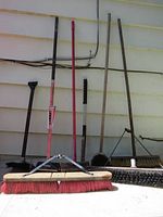 Photo showing 8 brooms and a metal rake leaning against the wall, includes used brooms and two contractor broom heads wrapped and unused.