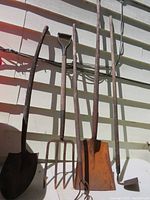 Lot of five garden tools including two shovels, two forks, and one hoe leaning against a wall outside in sunlight.