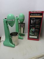 Two vintage green Hamilton Beach DrinkMaster mixers with stainless steel cups side by side with original box showing front panel.