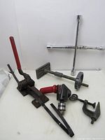 Photo of six man tools featuring a strapping tool, drill attachment, tire cross wrench, vintage clamp, rod end with flat piece, and wheel component.