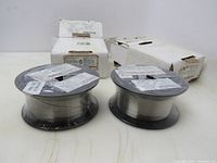 Two sealed spools of shiny stainless steel welding wire with original white boxes behind, wire diameter 0.030 inch (0.8 mm)