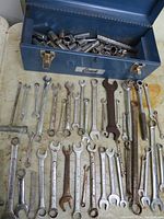 Metal toolbox with over 100 socket pieces inside and 41 assorted wrenches, including ratchets arranged in front of the box on a surface. Tools show used condition and some surface rust.