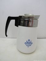 Side view of vintage coffee percolator showing white body with blue flowers, black handle and stainless steel top with glass knob lid.