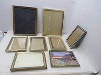 Eight vintage metal picture frames on a white surface, showing a variety of gold and silver tones with clear fronts and backing.