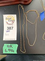 Two gold chain necklaces laid flat next to auction label '387' and handwritten '10K 6.94G' on green tape