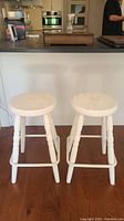 Front view of two white wooden bar stools placed on wood floor in front of counter.